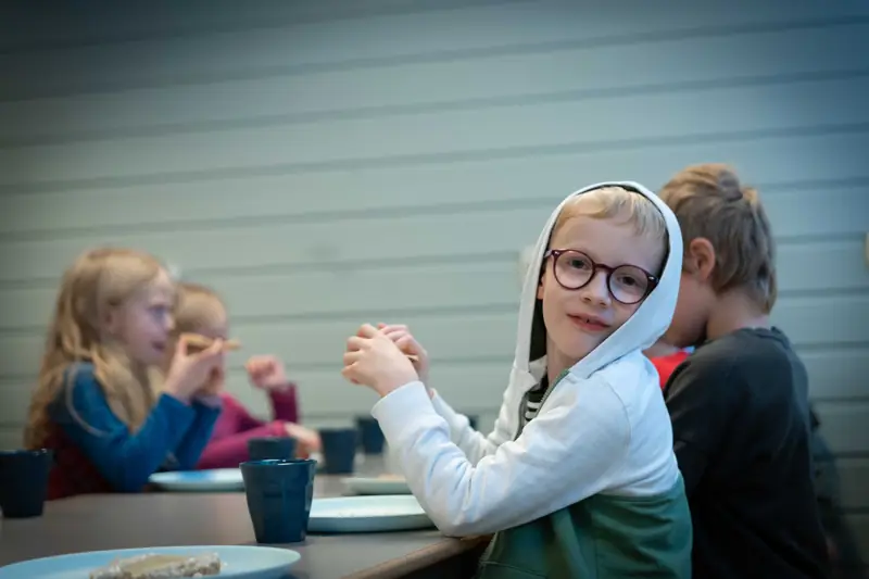 Smilende gutt i hettegenser ved lunsjbordet sammen med andre elever
